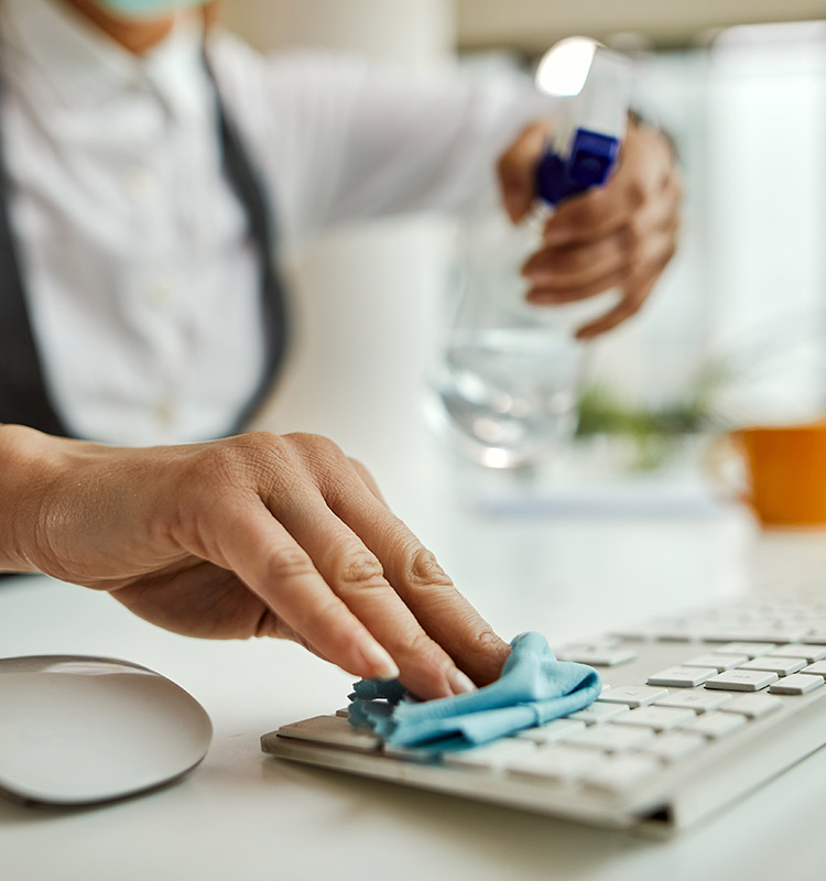 Person reinigt mit Sprühflasche und Putzlappen eine weiße Tastatur am Arbeitsplatz.