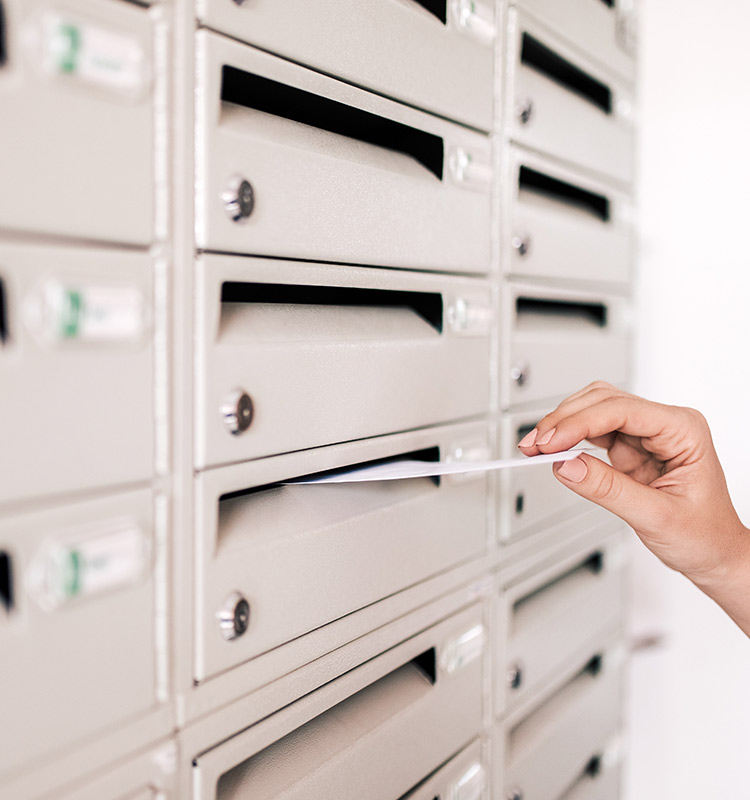Hand steckt Briefumschlag in weißes Briefkastensystem mit mehreren Postfächern.