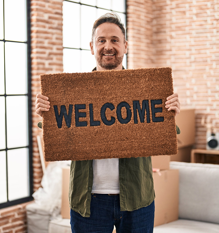 Mann hält eine Fußmatte aus Kokosfasern vor die Brust. Auf der Matte steht "Welcome".
